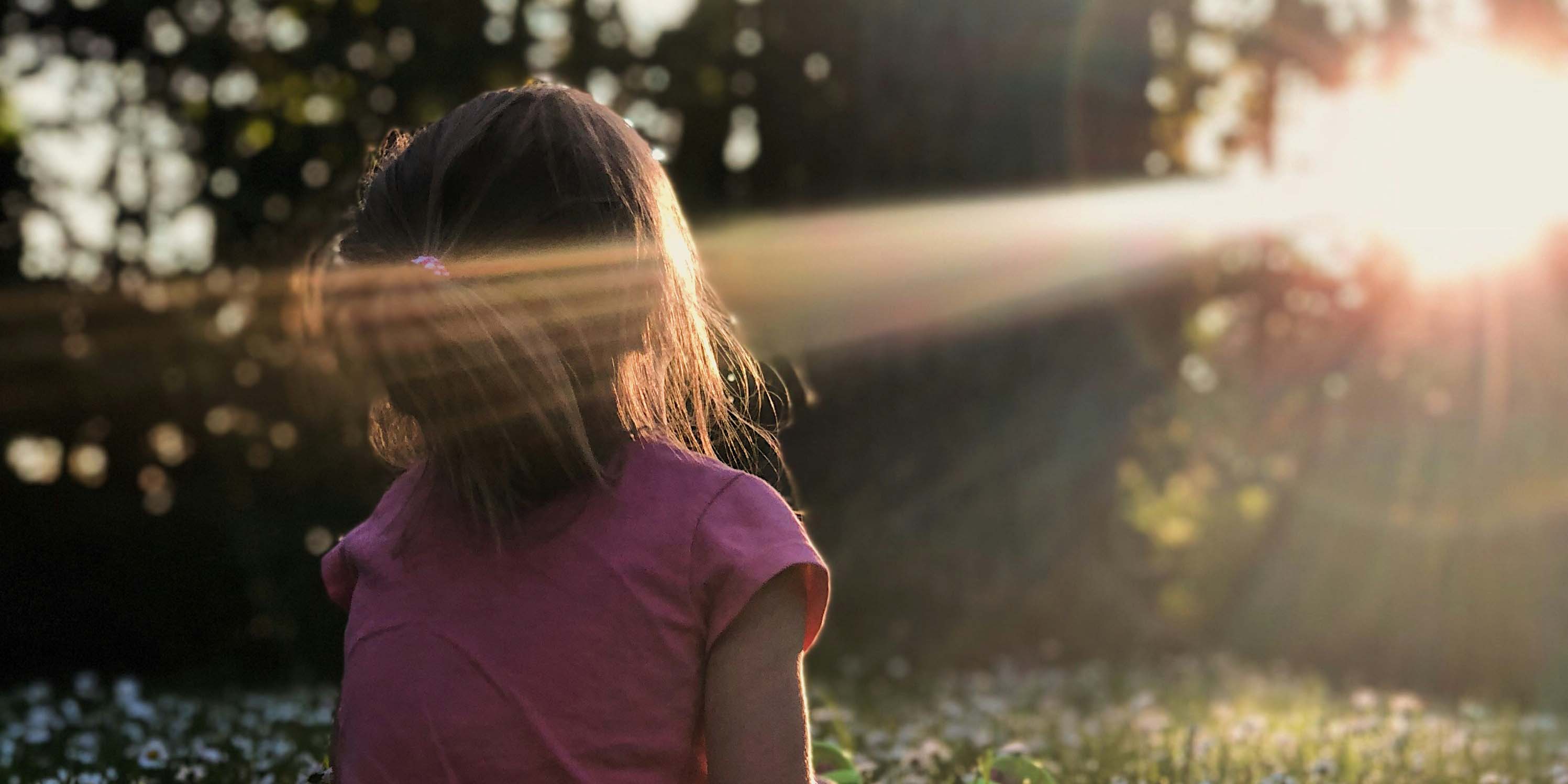 Auf dem Foto ist ein Mädchen von hinten zu sehen, das sich auf einer Waldlichtung befindet und von Sonnenstrahlen angeschienen wird.