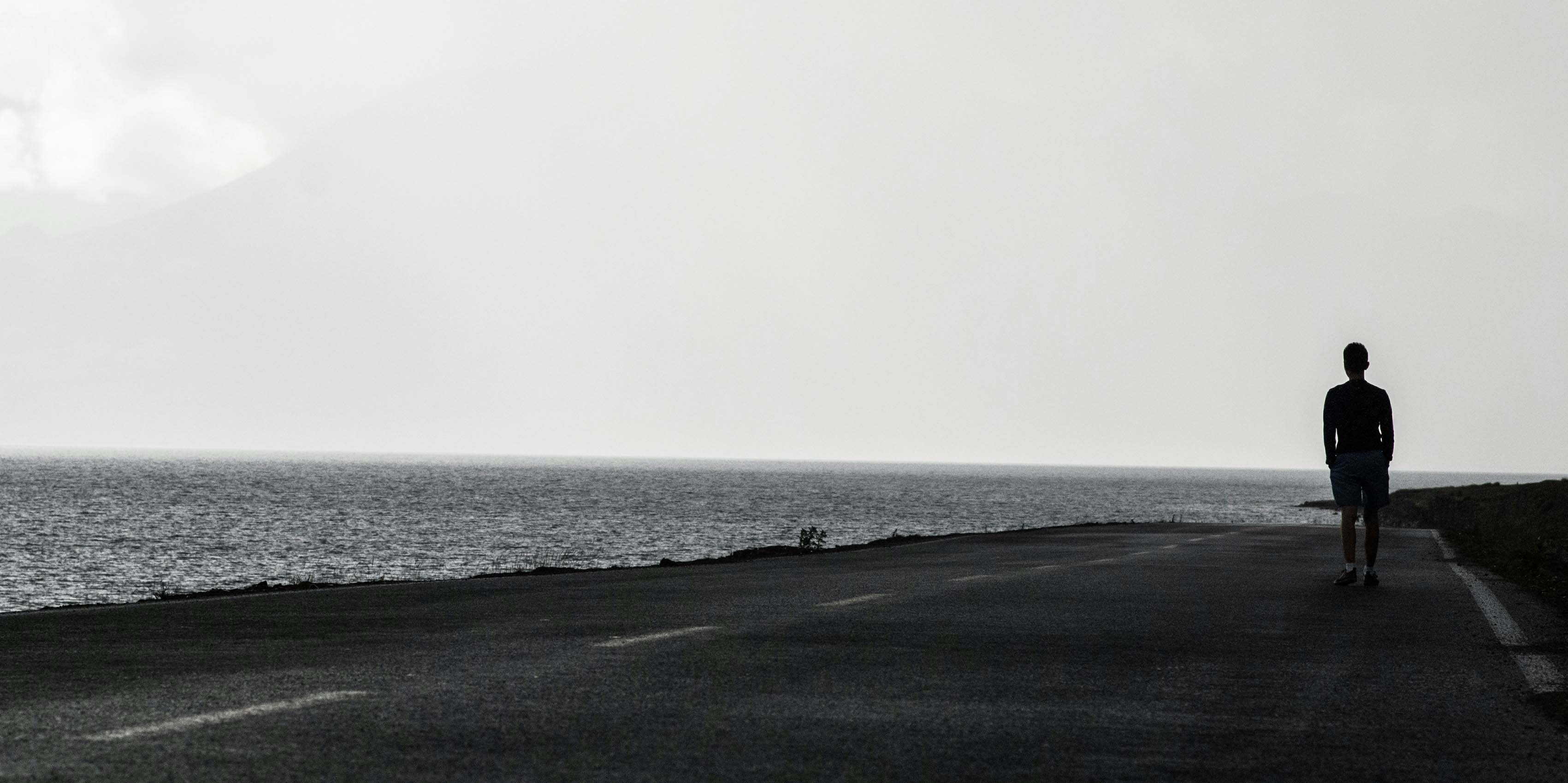 Auf dem Bild ist ein Mann auf einer einsamen Straße zu sehen, links neben sich das Meer.